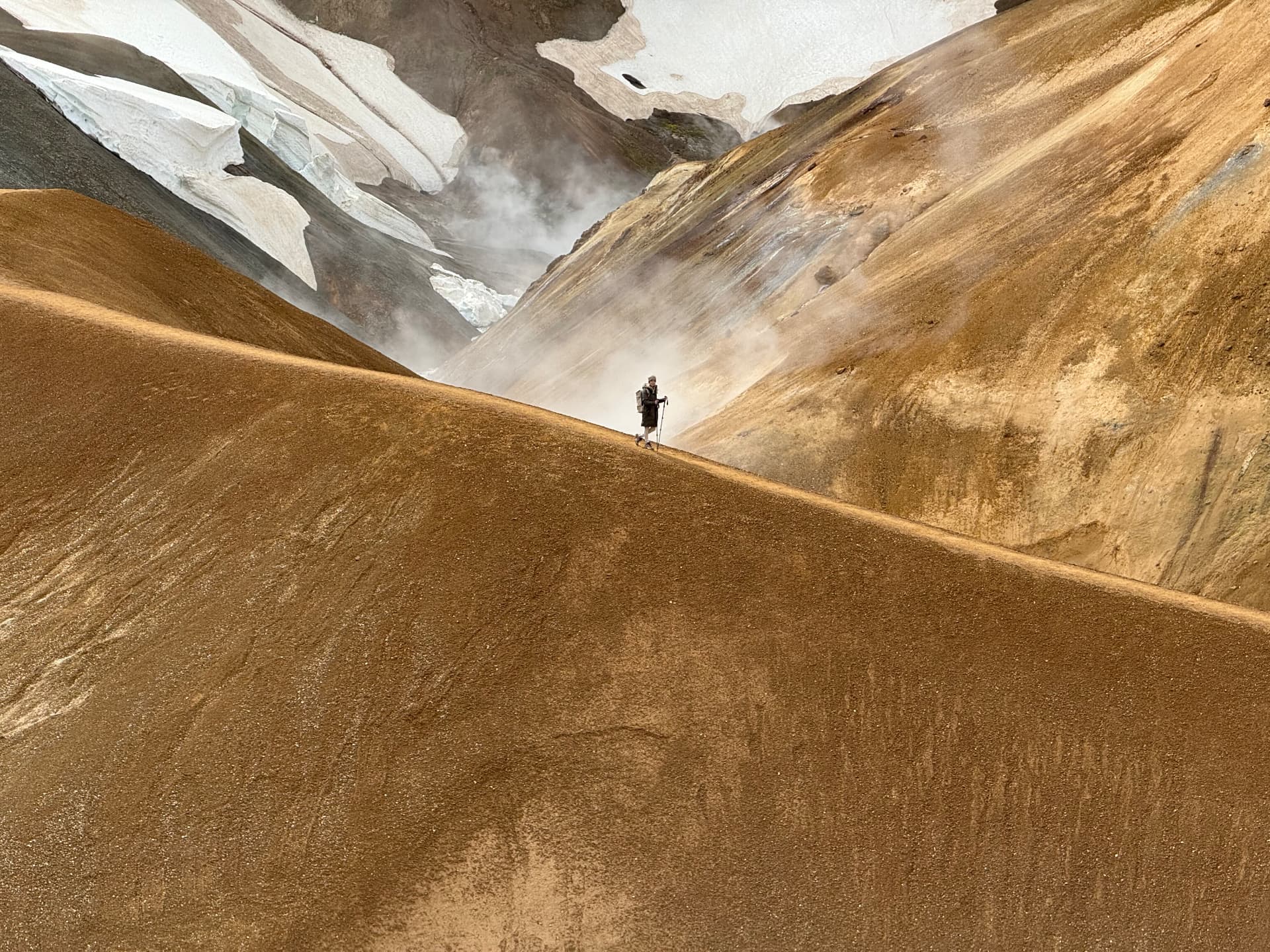A lone hiker crossing ochre volcanic ridges beside snow and steam in the Icelandic highlands.